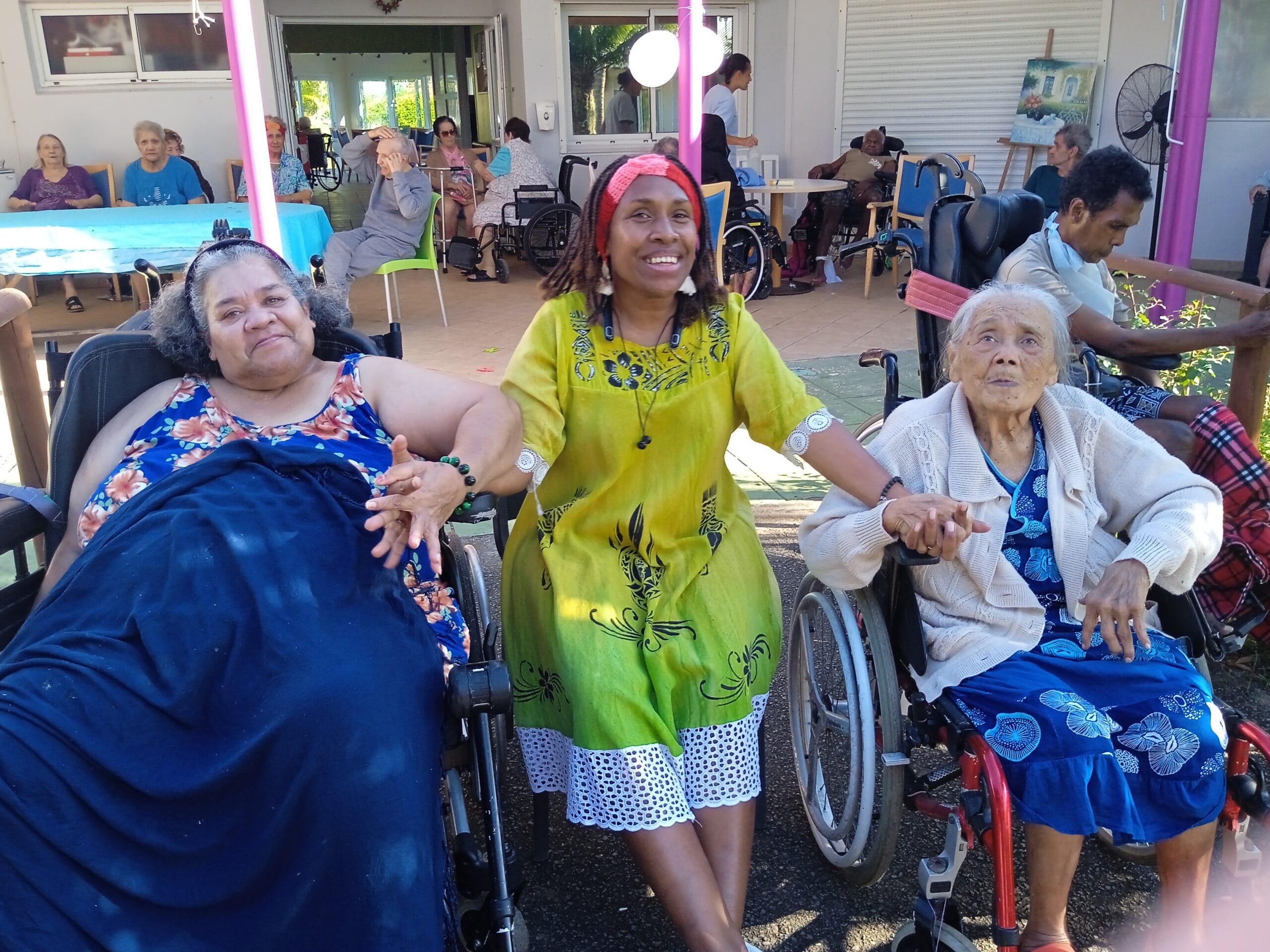 Trois résidentes de la Résidence La Cordyline souriantes et se tenant par la main, assises en plein air lors d'une activité conviviale